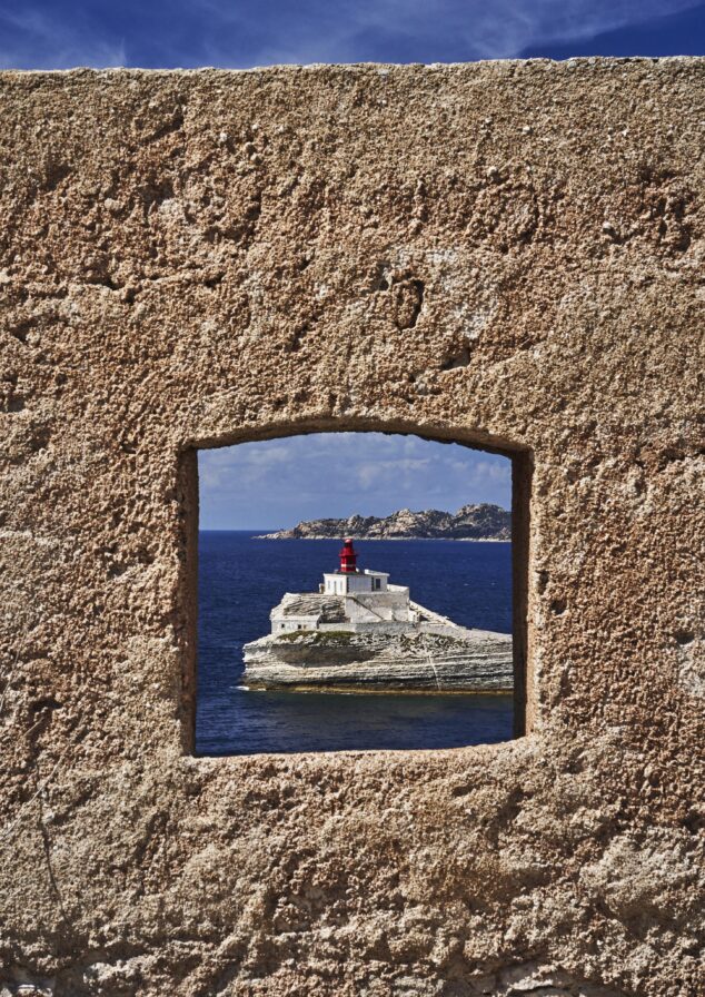 Lighthouse on rocky island seen through a square stone window. Blue sea and partially cloudy sky in the background.