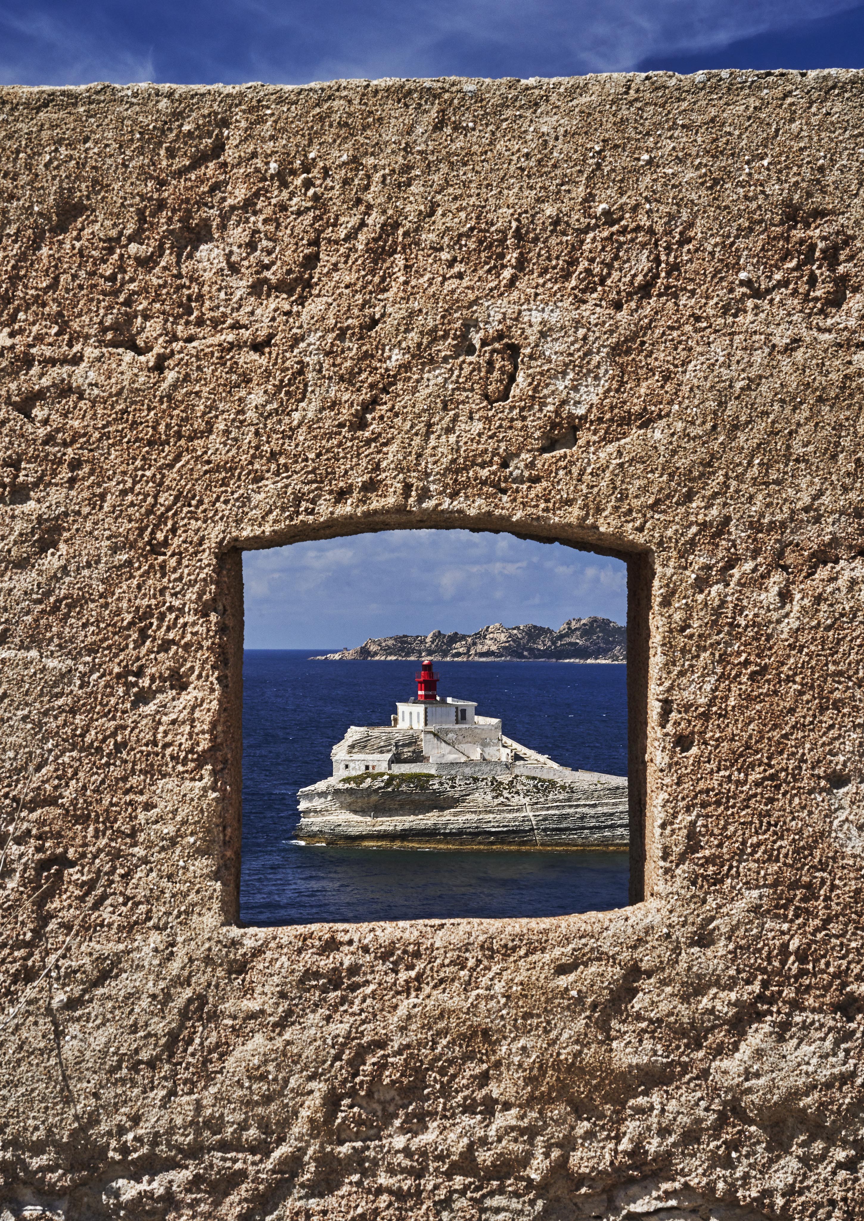 Lighthouse on rocky island seen through a square stone window. Blue sea and partially cloudy sky in the background.