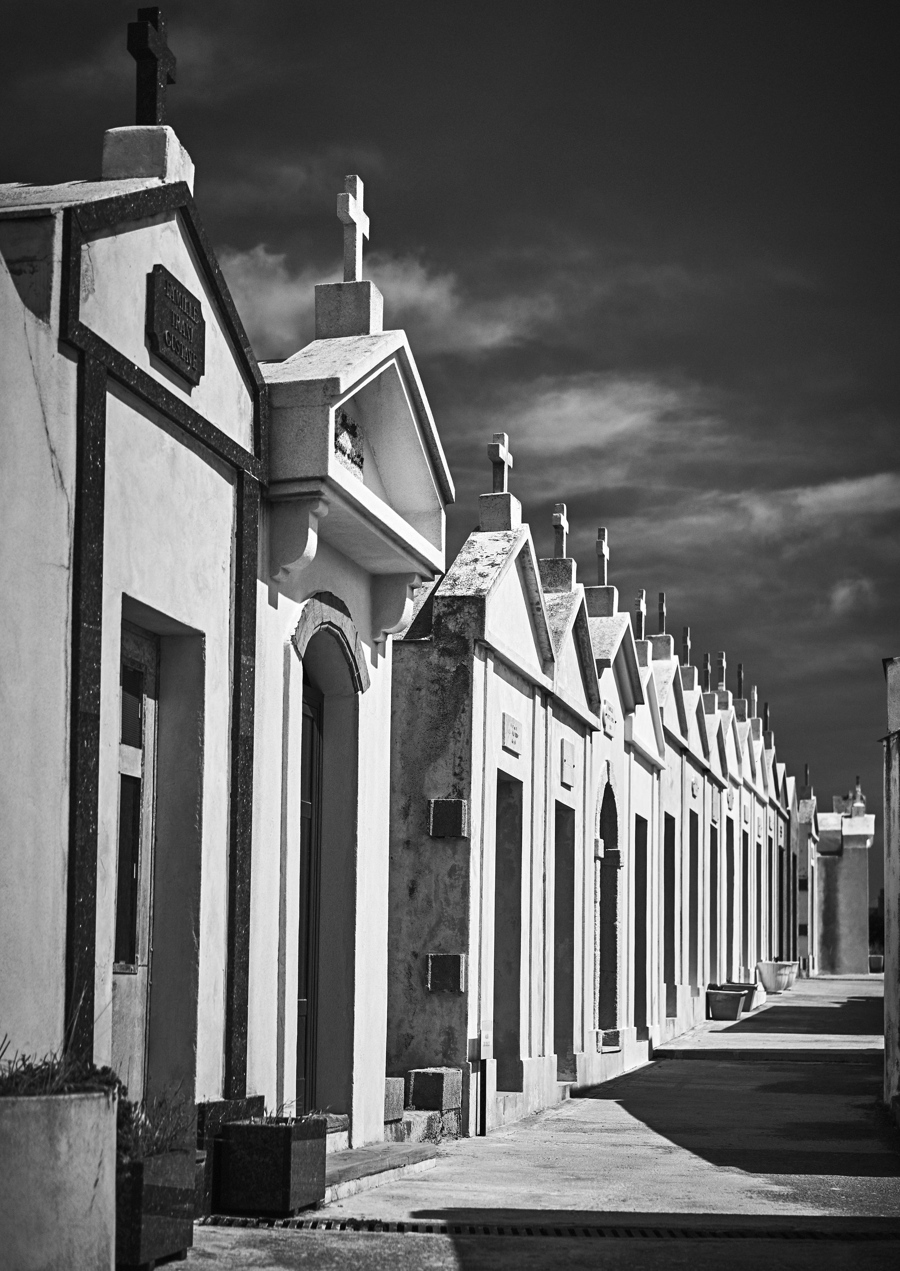 Row of ornate above-ground tombs with crosses in a cemetery under a cloudy sky, captured in black and white.