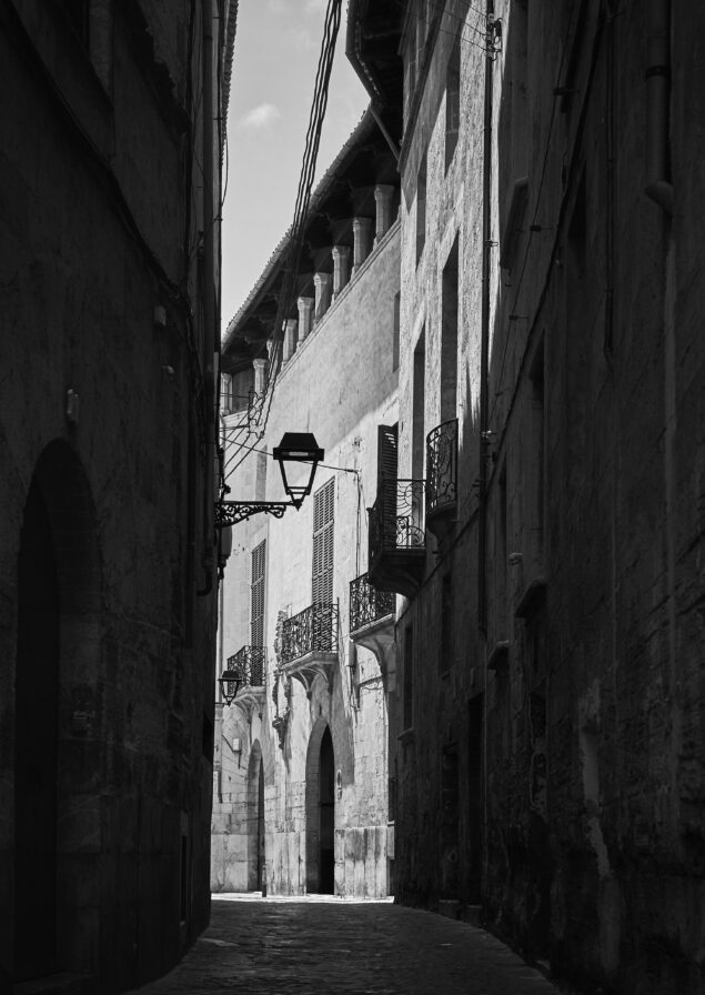 Narrow alleyway between tall, historic buildings with decorative balconies and a single street lamp, illuminated by sunlight.