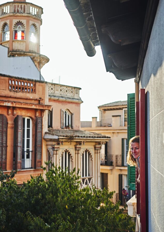 Person peeking out of a window in a building, with an ornate tower and surrounding structures visible in the background.