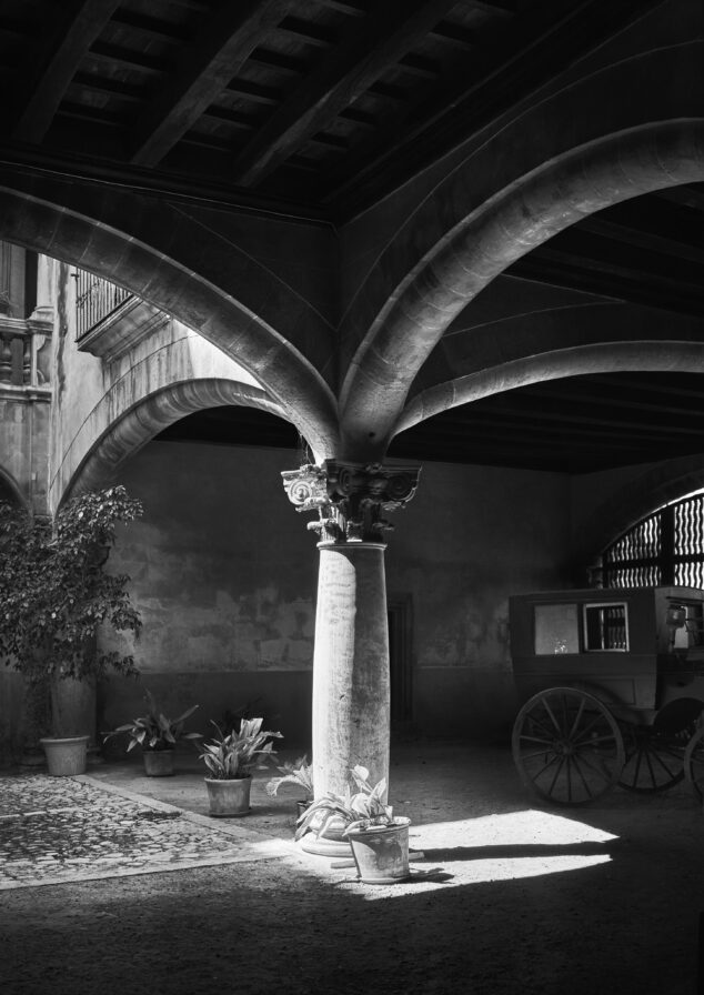 A black-and-white image of an old stone courtyard with a central column supporting arches. Potted plants adorn the base, and a wooden cart is partially visible in the background.