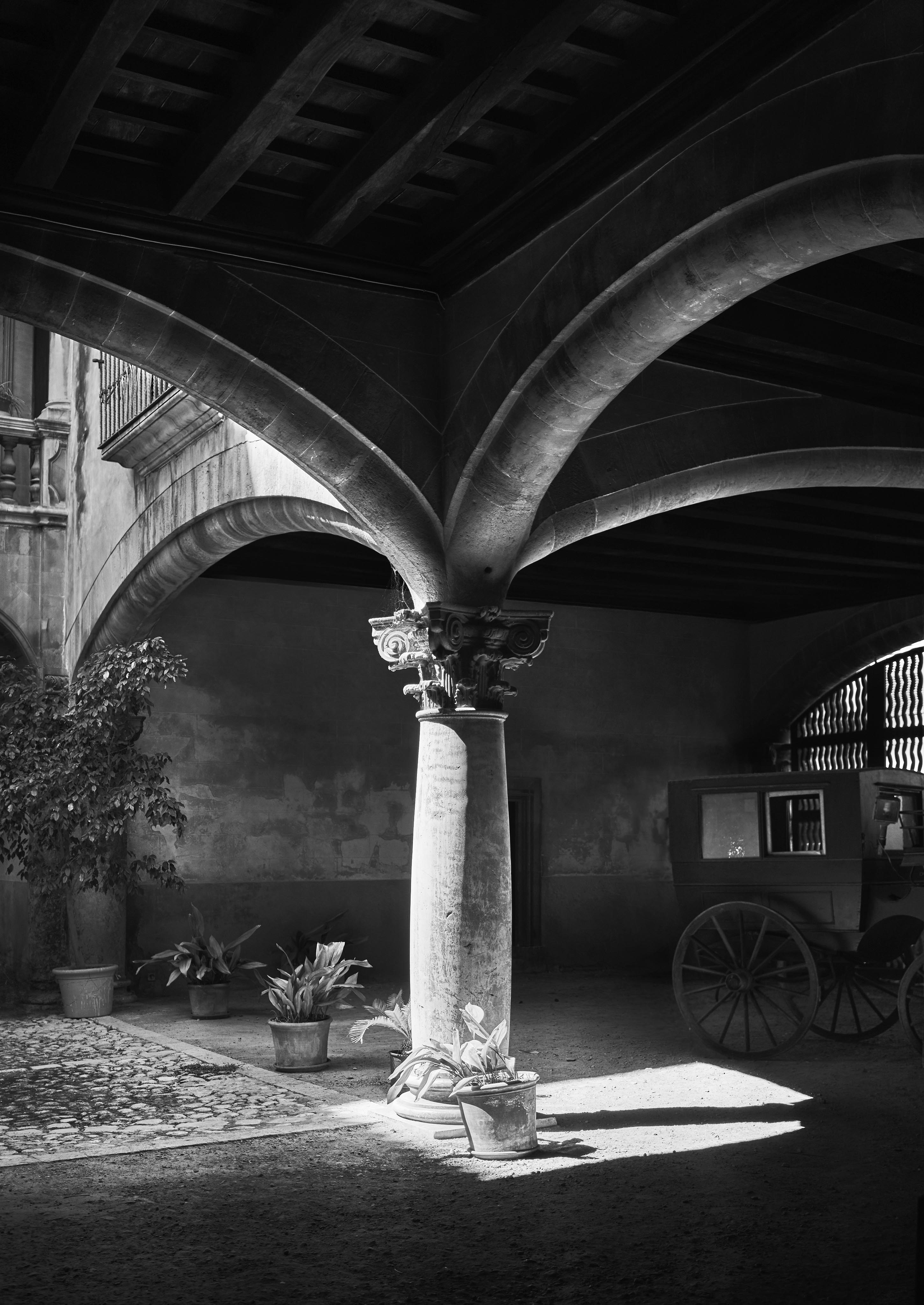 A black-and-white image of an old stone courtyard with a central column supporting arches. Potted plants adorn the base, and a wooden cart is partially visible in the background.