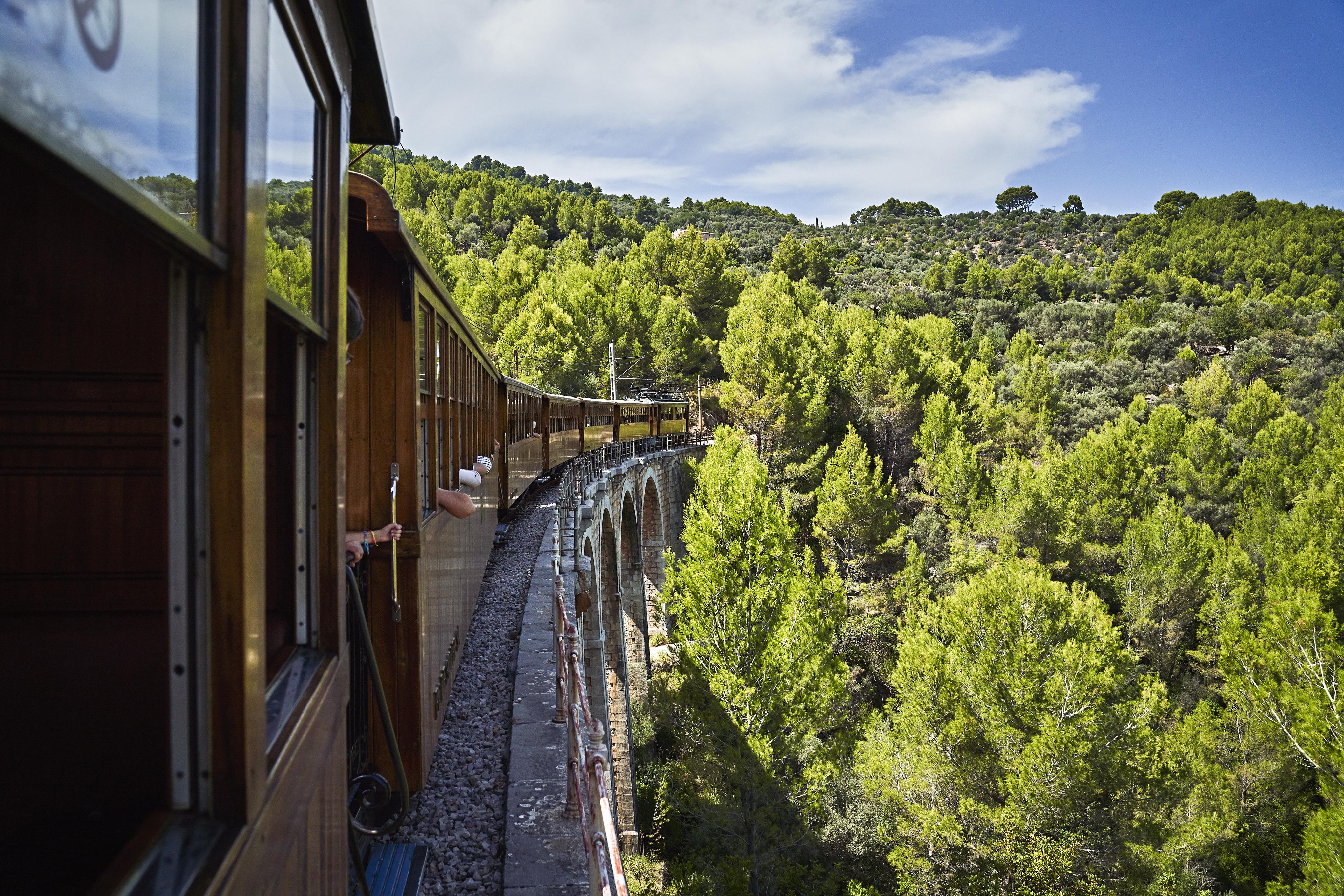 A vintage train travels across a curved stone bridge surrounded by dense green forest and under a partly cloudy sky. Passengers’ arms are visible leaning out of the windows.