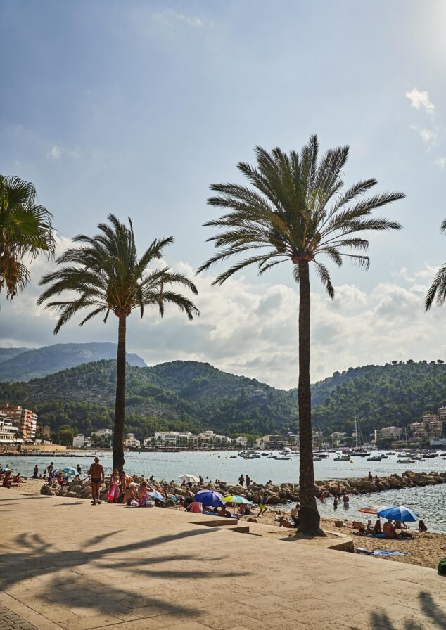 Beach scene with people sunbathing and relaxing near palm trees. Sailboats are on the water, and mountains are visible in the background under a blue sky with clouds.