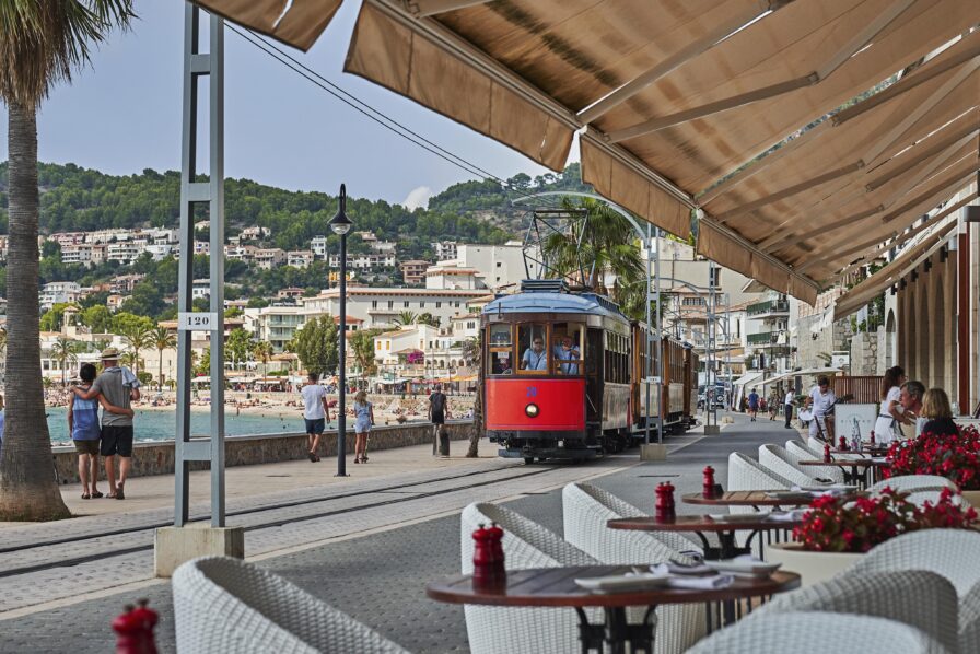 A red and blue tram travels along the waterfront promenade near a beach. Outdoor cafes with white chairs line the street under large awnings. Hills with buildings are visible in the background.
