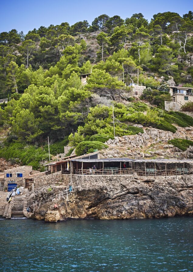 A rustic seaside building with a terrace is surrounded by lush greenery and rocky terrain, facing a calm blue sea. Steps lead down to the water.