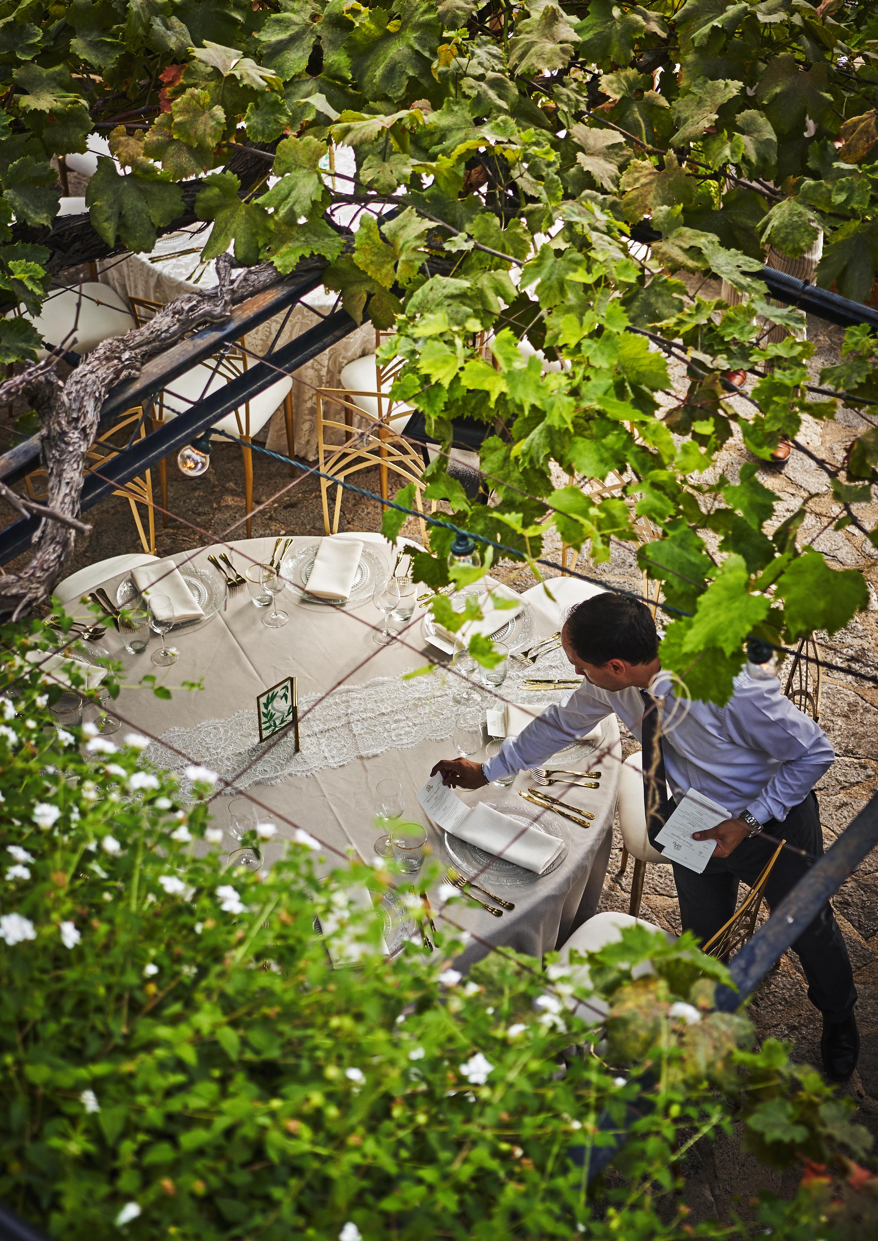 A waiter sets an outdoor dining table with white linens and tableware under a vine-covered canopy.