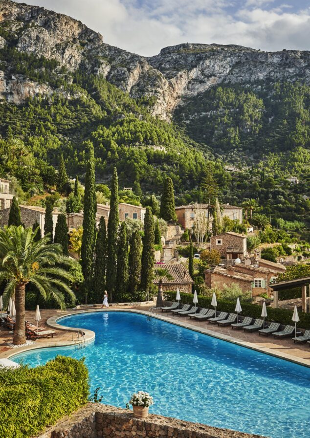 A large, clear blue pool surrounded by deck chairs and greenery, set against a backdrop of mountains and quaint stone buildings under a partly cloudy sky.