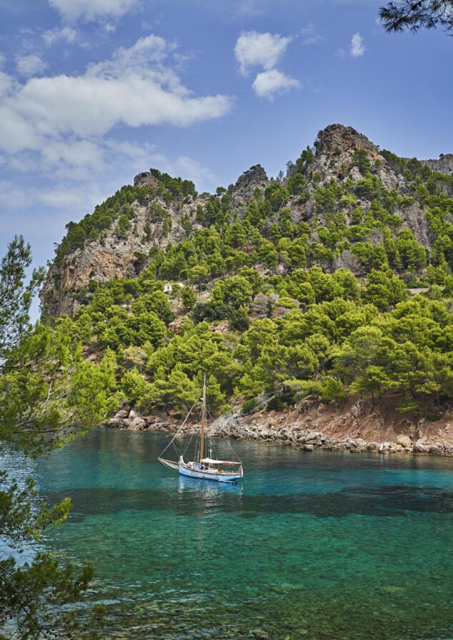 A sailboat is anchored in a clear, turquoise bay surrounded by rocky cliffs and dense green pine trees under a partly cloudy sky.