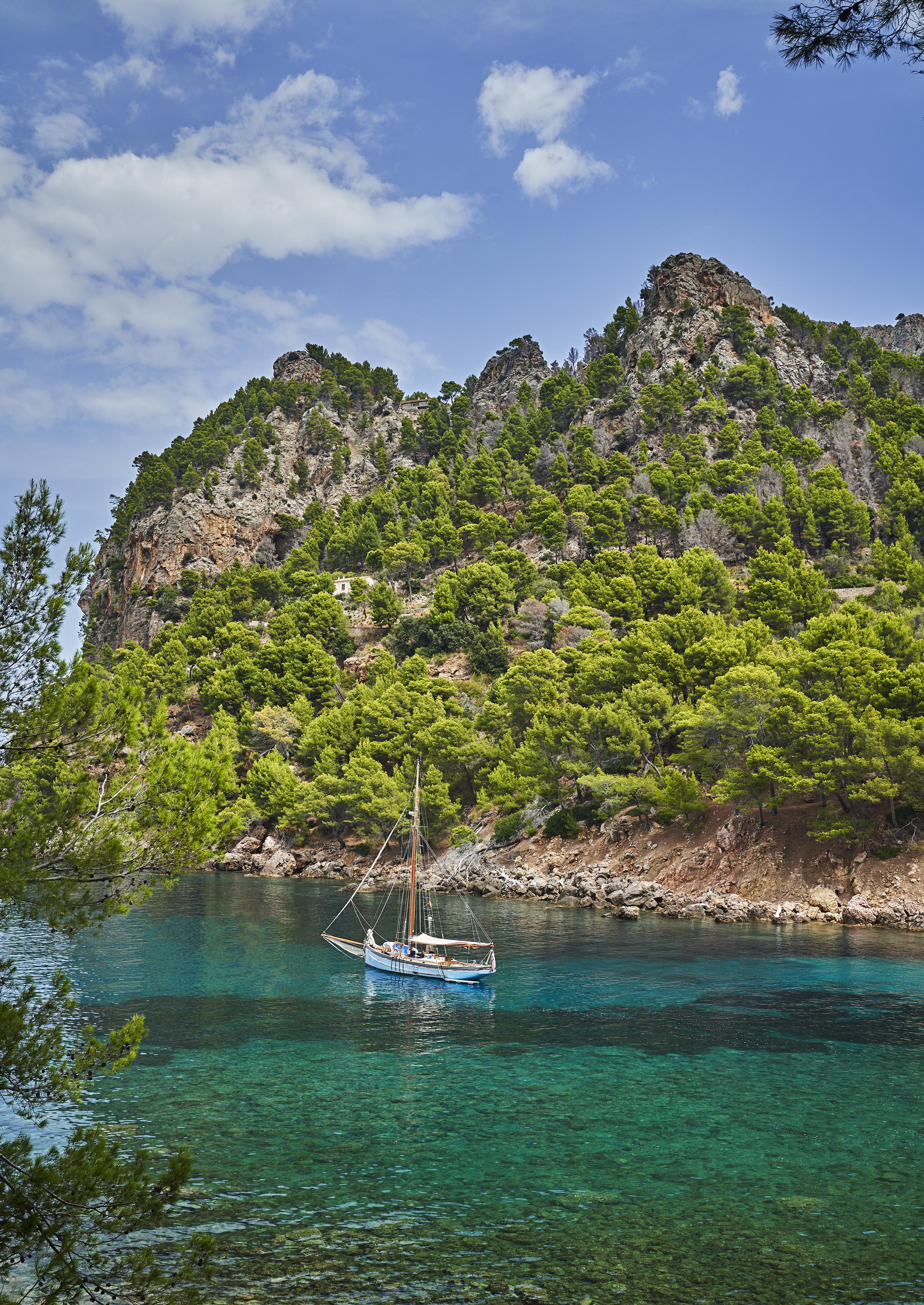 A sailboat is anchored in a clear, turquoise bay surrounded by rocky cliffs and dense green pine trees under a partly cloudy sky.