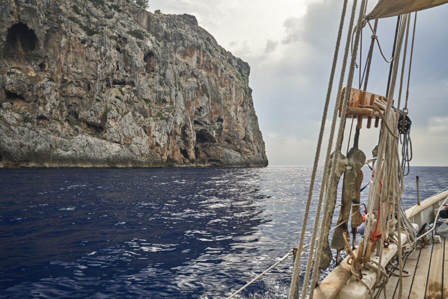 Sailing boat near a rocky cliff on a calm sea under a cloudy sky.