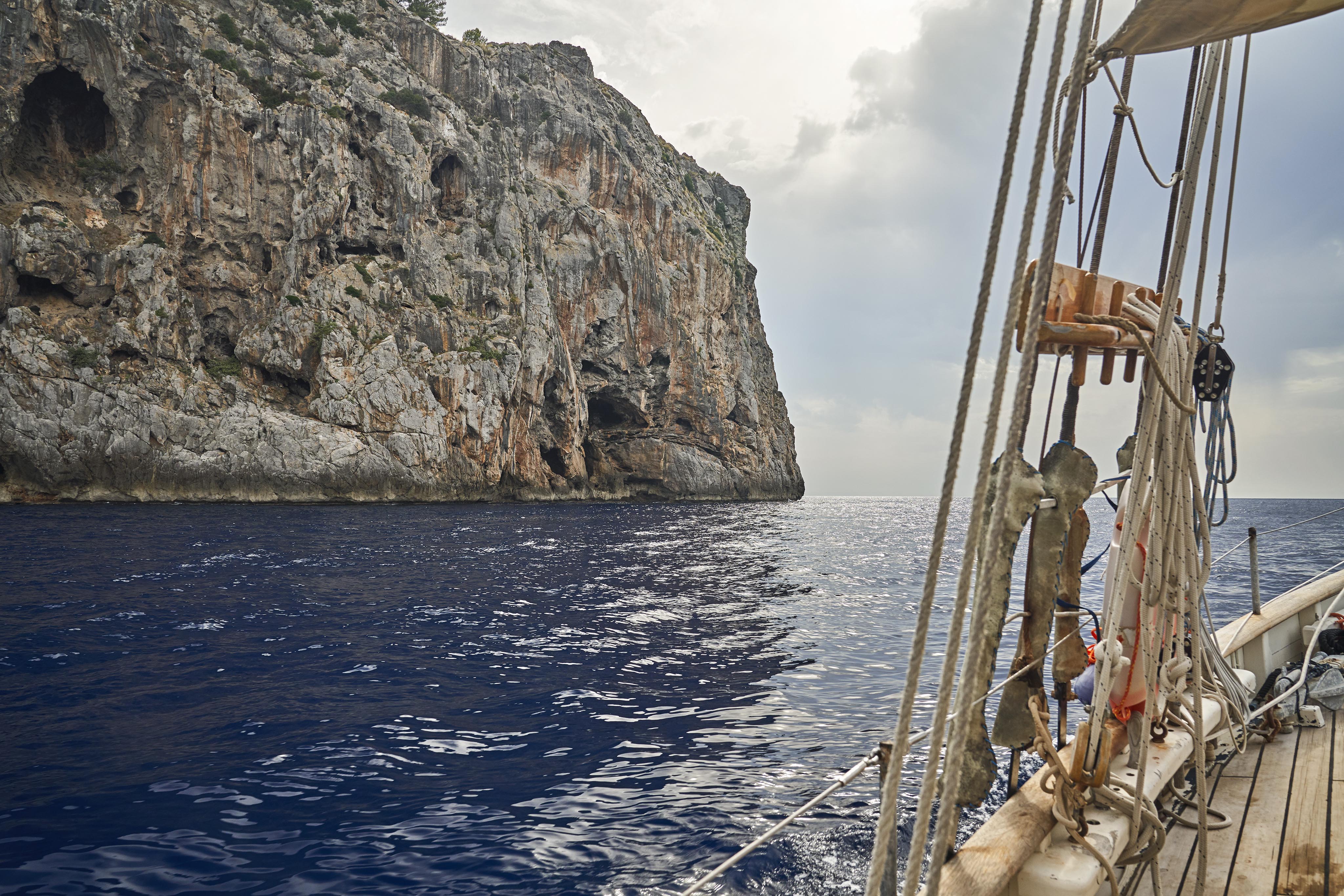 Sailing boat near a rocky cliff on a calm sea under a cloudy sky.
