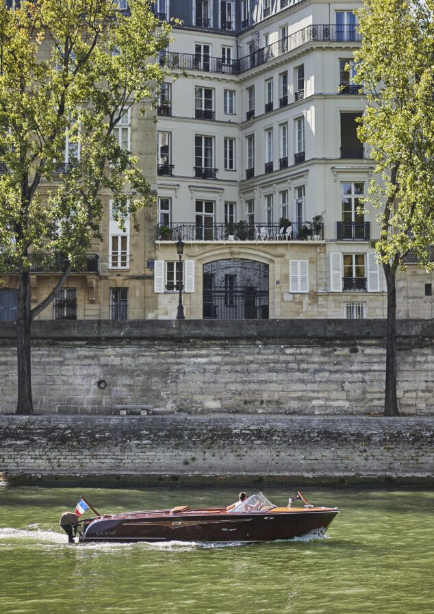 A wooden motorboat travels along a river, with a multi-story building and trees visible in the background.