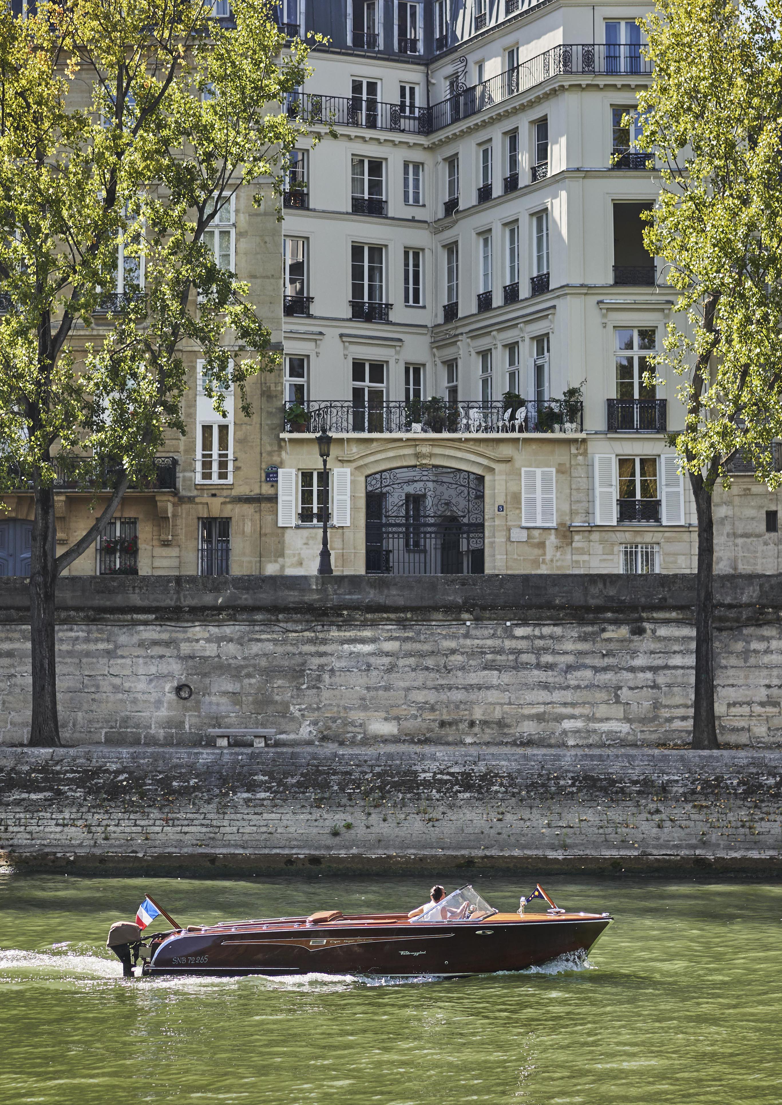 A wooden motorboat travels along a river, with a multi-story building and trees visible in the background.