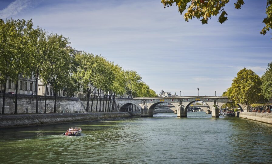 A stone bridge spans a calm river in a leafy urban area, with a small boat passing beneath and trees lining the banks under a partly cloudy sky.