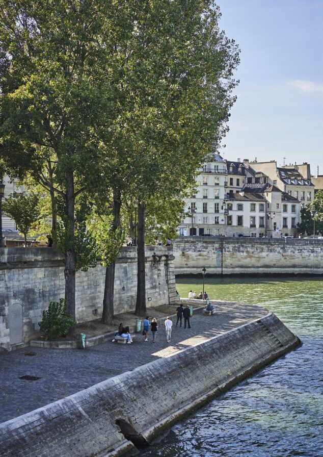 People walk and sit by the river under tall trees on a cobblestone path, with historic buildings in the background.