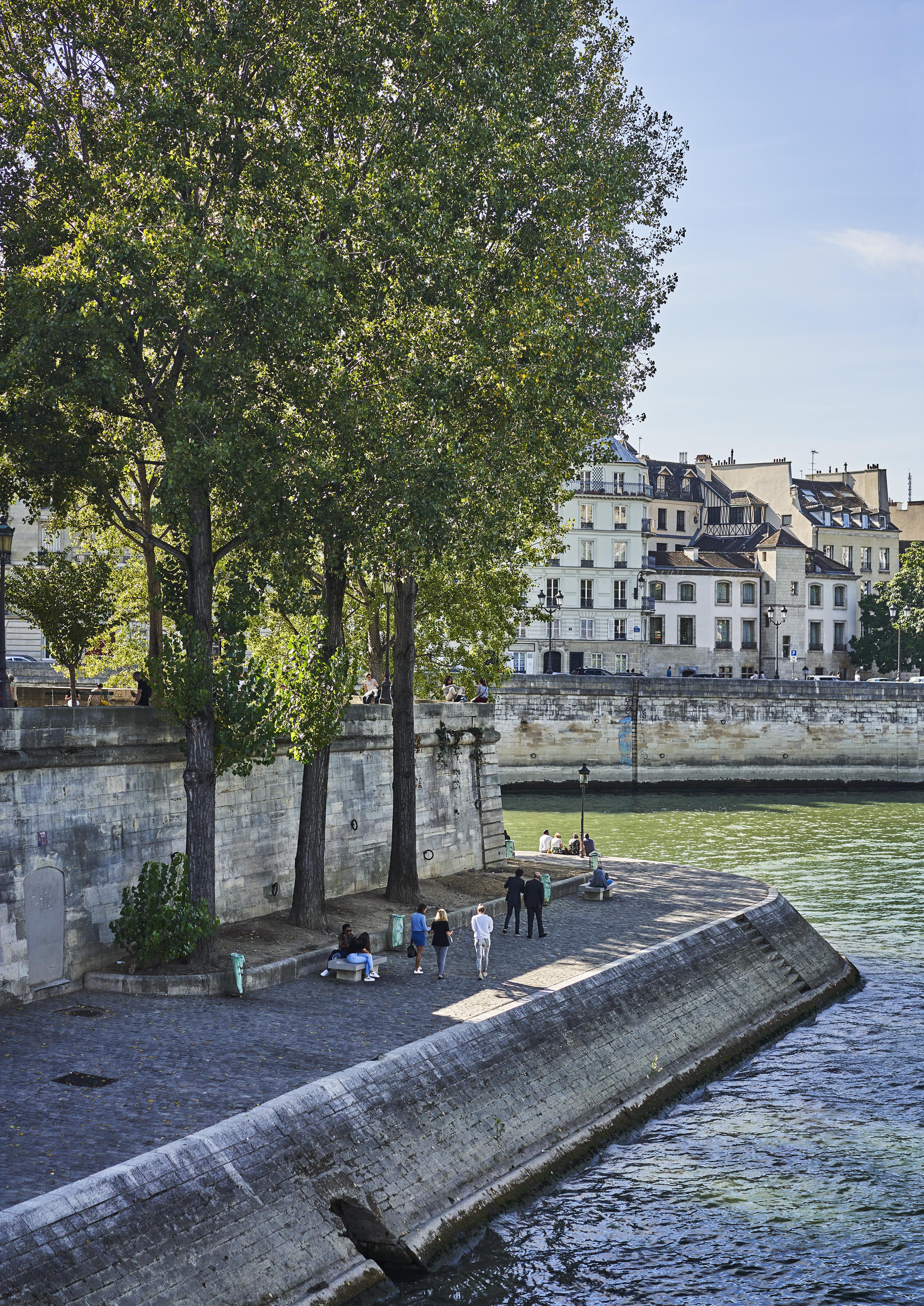 People walk and sit by the river under tall trees on a cobblestone path, with historic buildings in the background.