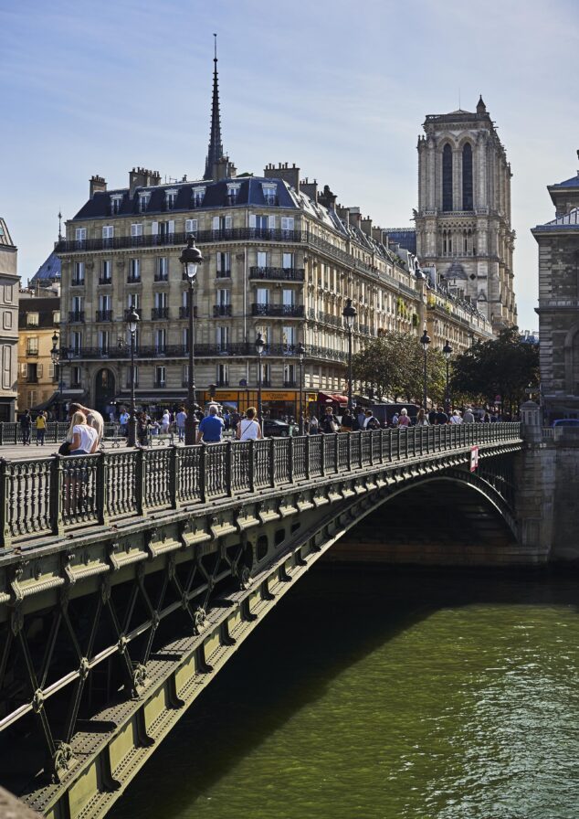 A bridge with pedestrians crosses a river in Paris, with ornate buildings and a tower in the background on a clear day.