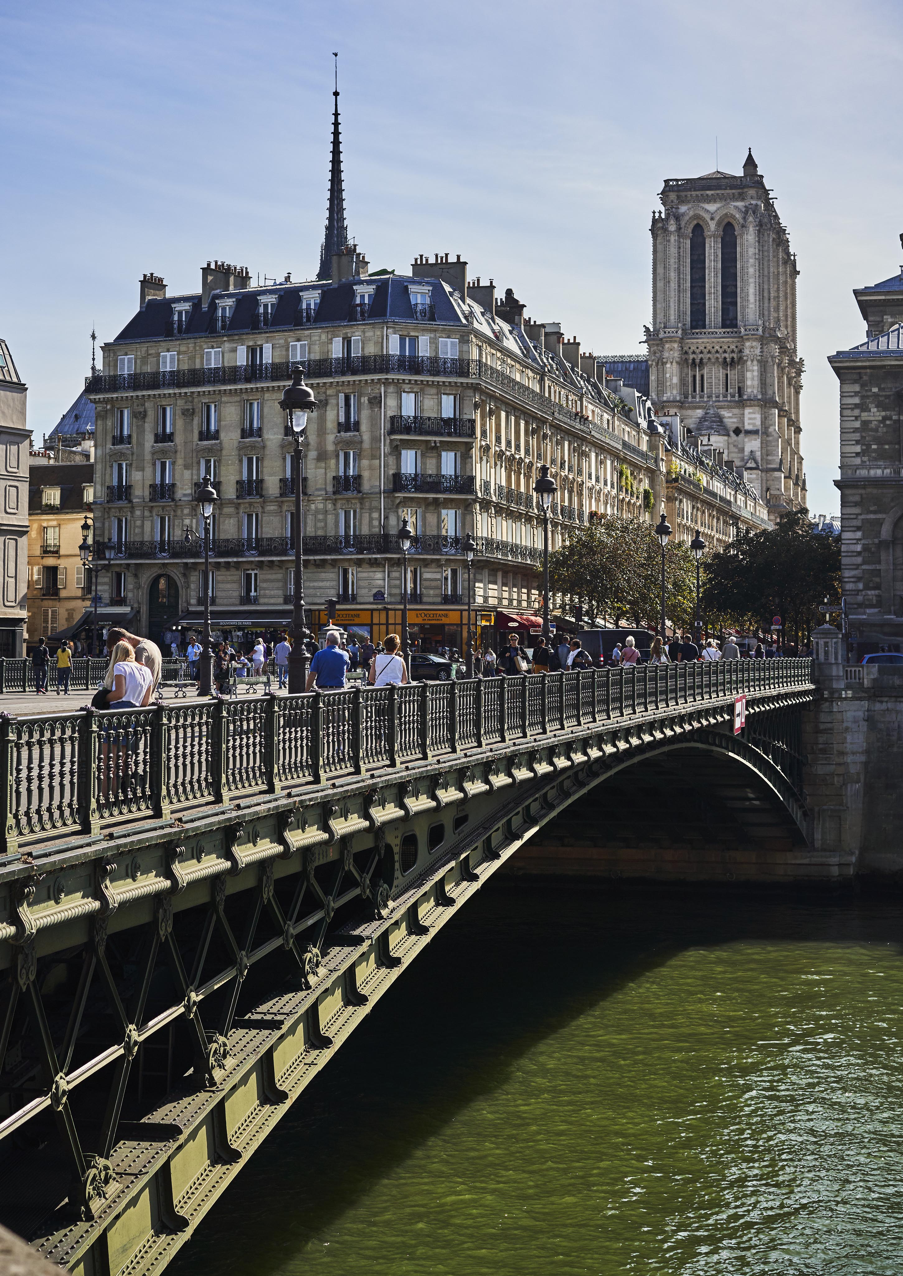 A bridge with pedestrians crosses a river in Paris, with ornate buildings and a tower in the background on a clear day.