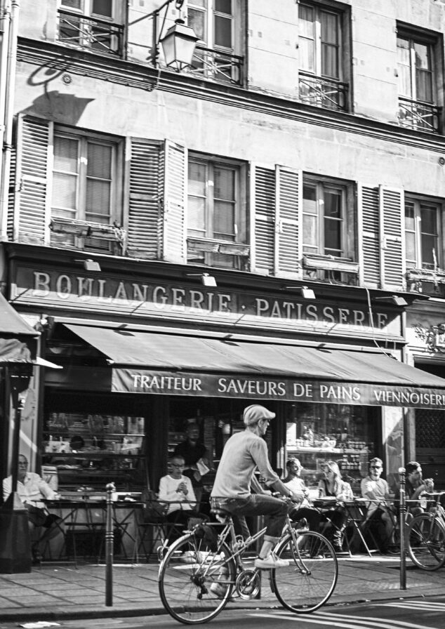 A person rides a bicycle past a boulangerie-patisserie in a city. People are seated outside the bakery, and the building has multiple floors with windows. Black and white image.