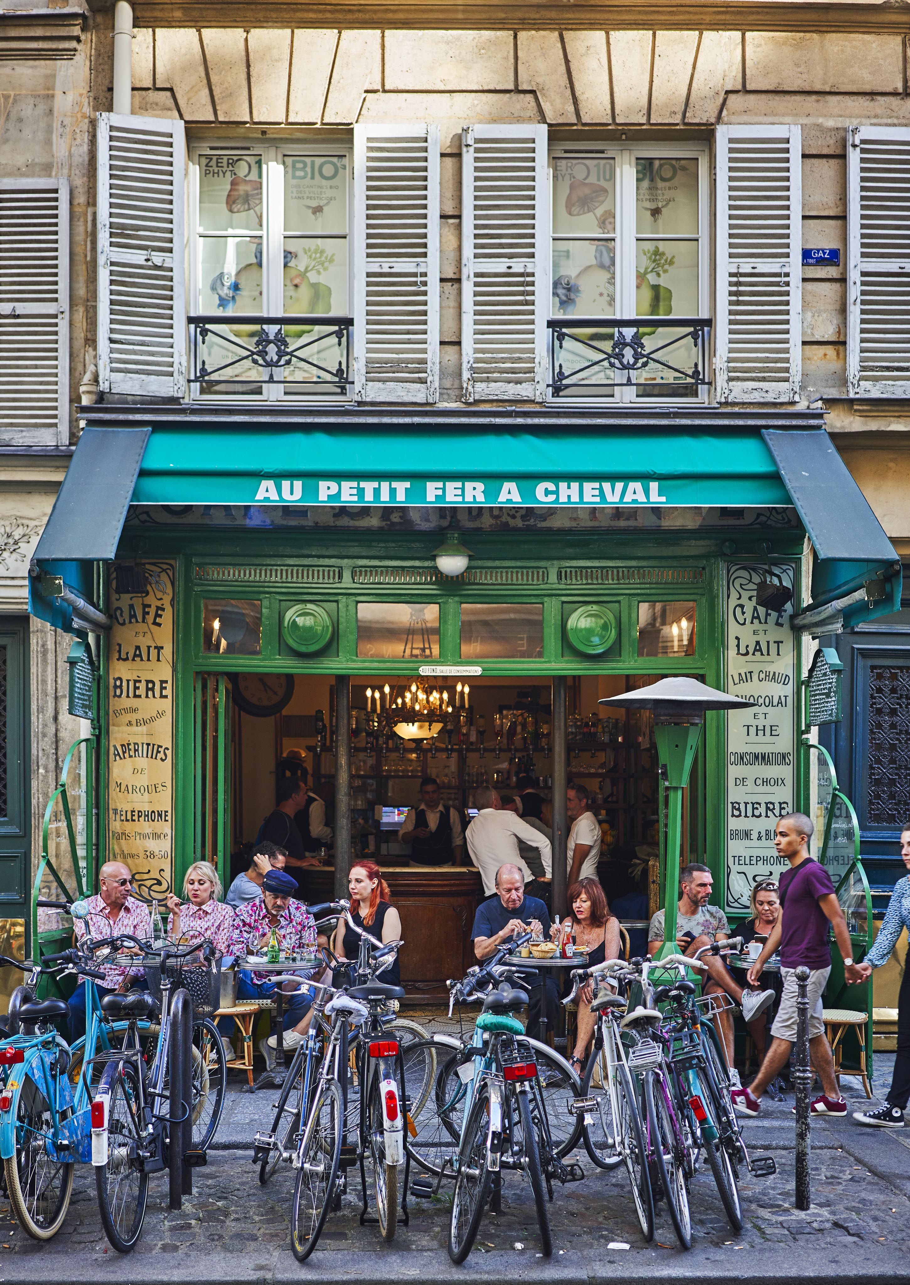 People sitting at outdoor tables of a café with green awning. Several bicycles are parked in front. Café facade features wooden details and signage displaying menu items.