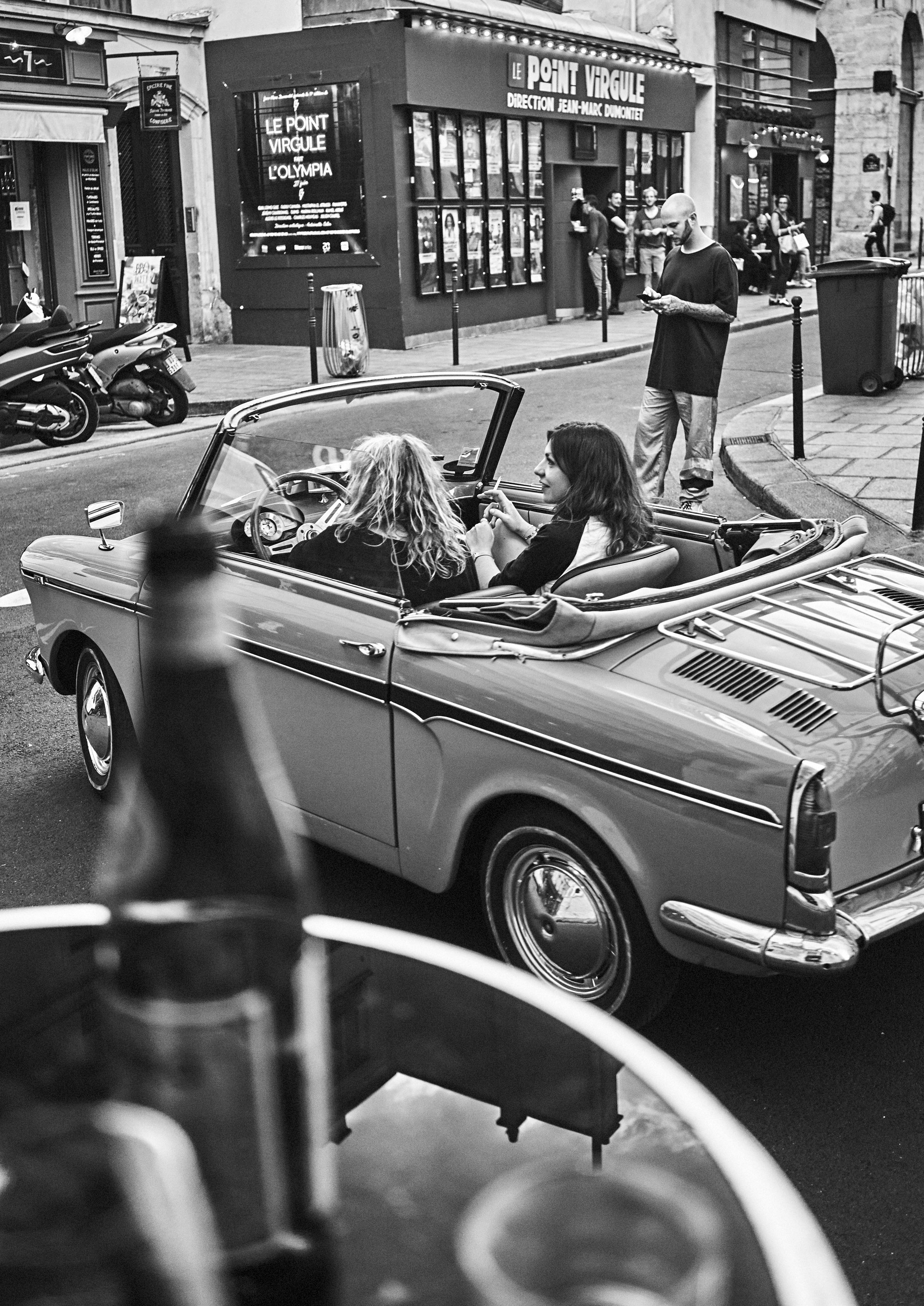 Two women sit in a parked convertible on a street. A man stands nearby holding a drink. A blurred bottle is in the foreground. Building signs are visible in the background.