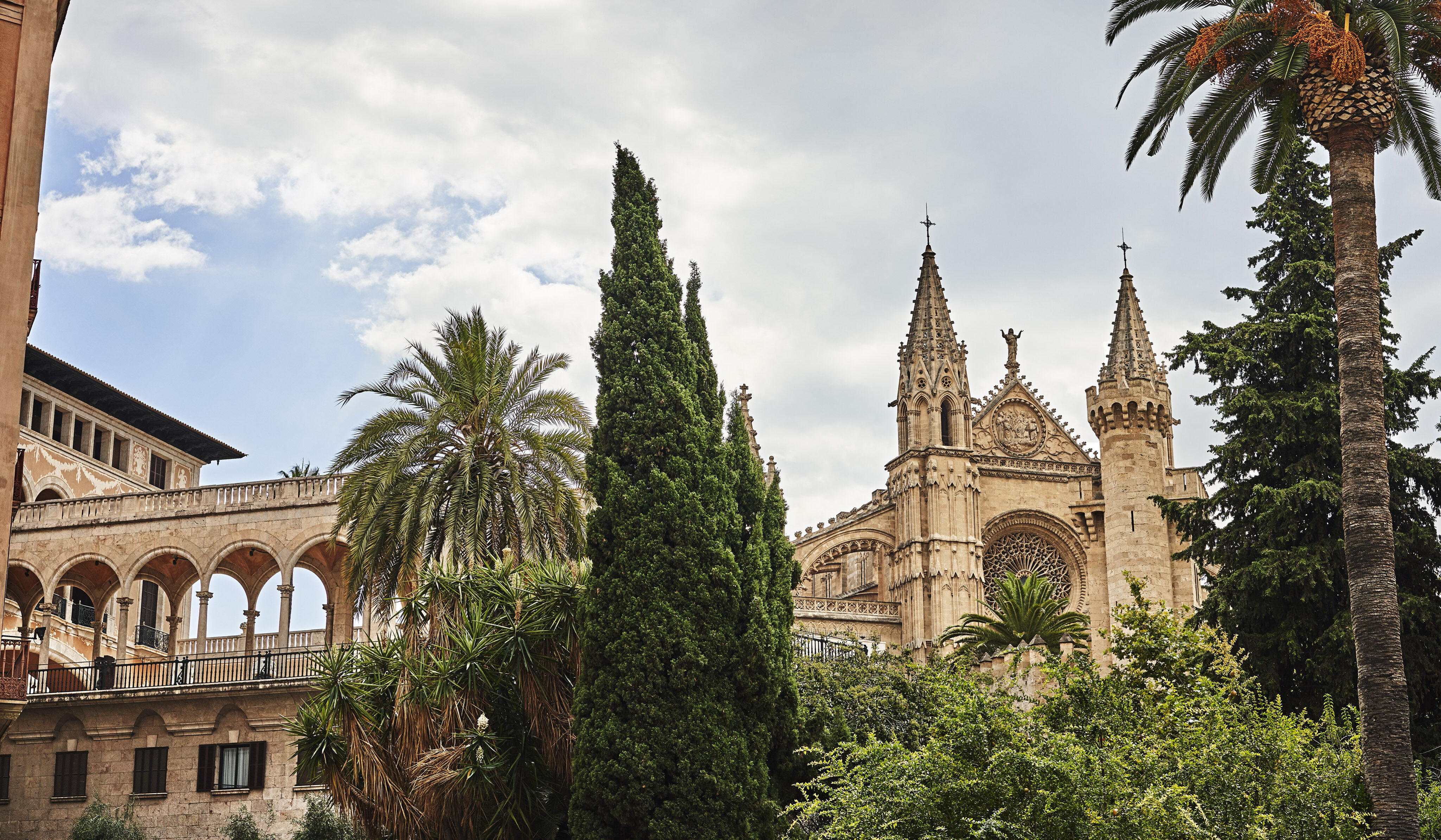 Gothic cathedral with ornate spires in a lush garden setting, featuring palm and cypress trees. Arched walkway is visible, with a cloudy sky above.