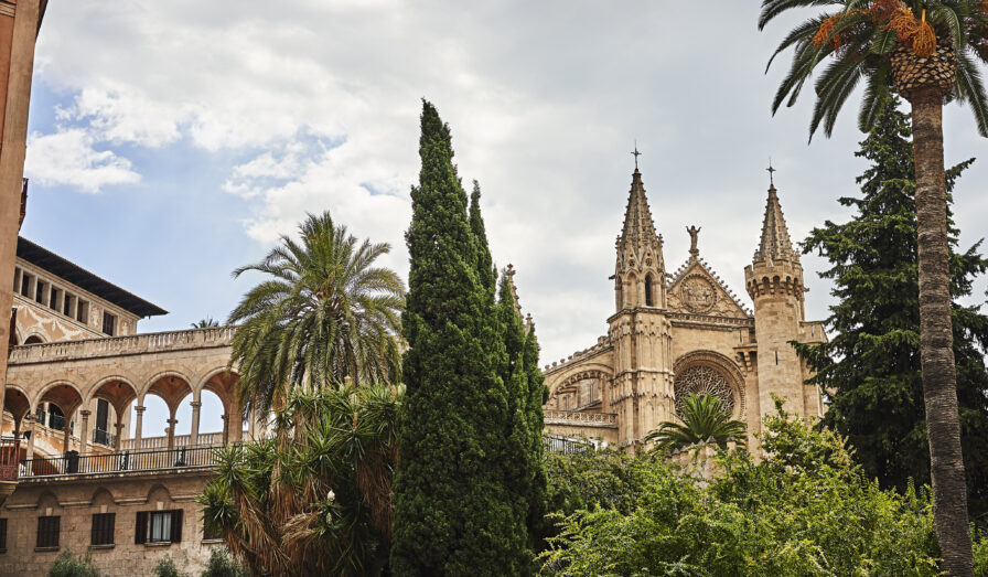 Gothic cathedral with ornate spires in a lush garden setting, featuring palm and cypress trees. Arched walkway is visible, with a cloudy sky above.