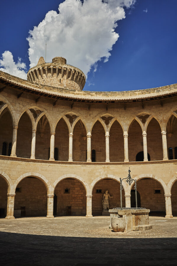 Round stone courtyard with arched colonnade and central well, beneath a blue sky with clouds. Ancient stone tower rises above the arches.