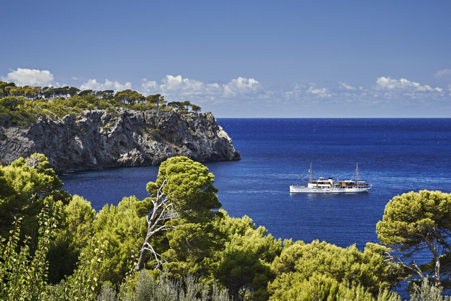 A white boat is on a calm blue sea near a rocky coastline with trees under a clear blue sky.