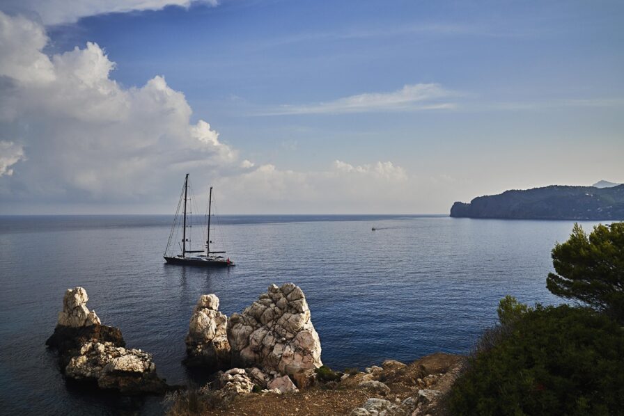 Sailboat on calm Mallorca sea near rocky Deia coast under a partly cloudy sky, with distant cliffs and greenery.
