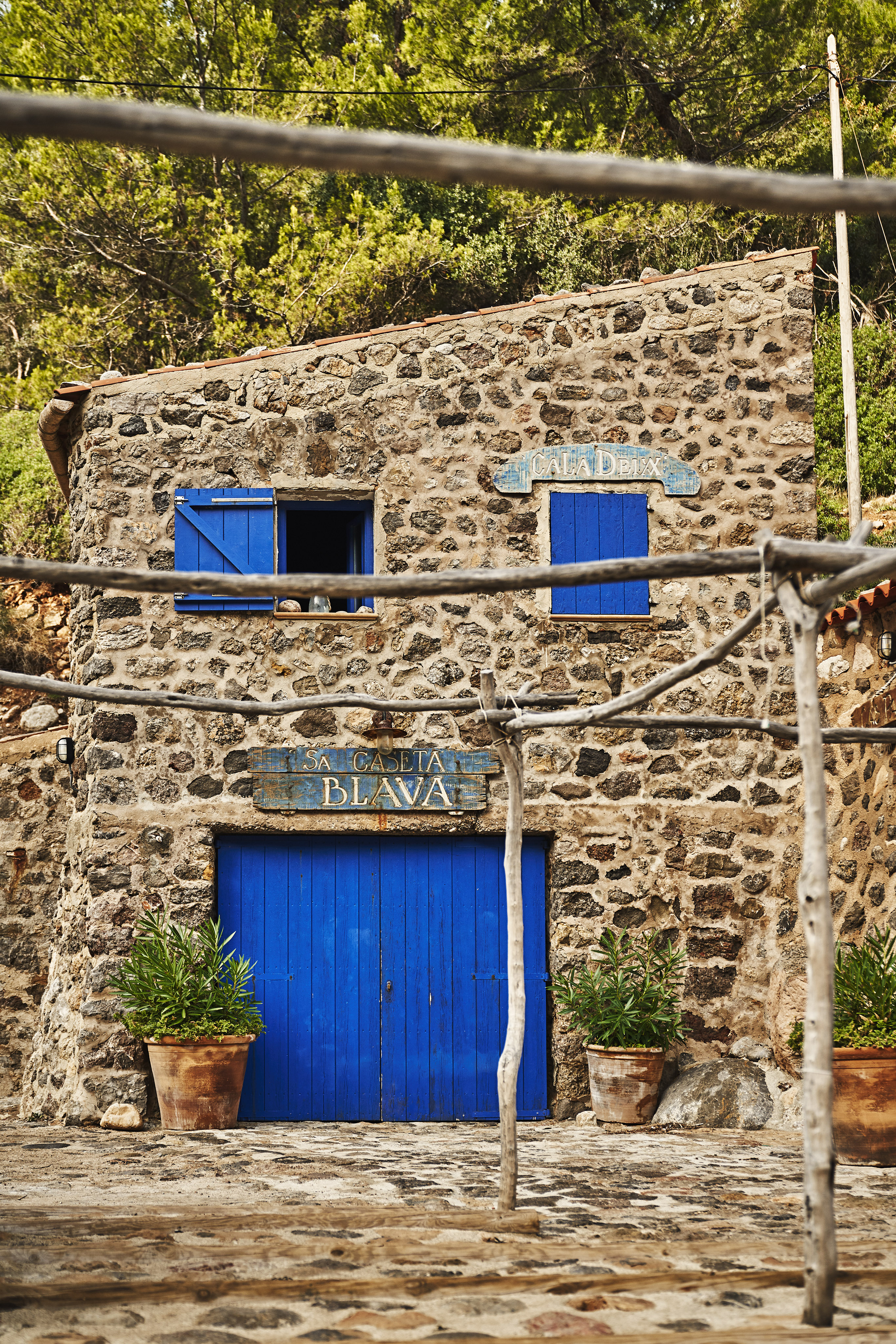 Stone building with bright blue doors and shutters, surrounded by trees. Two potted plants flank the entrance. Wooden framework is visible in the foreground.