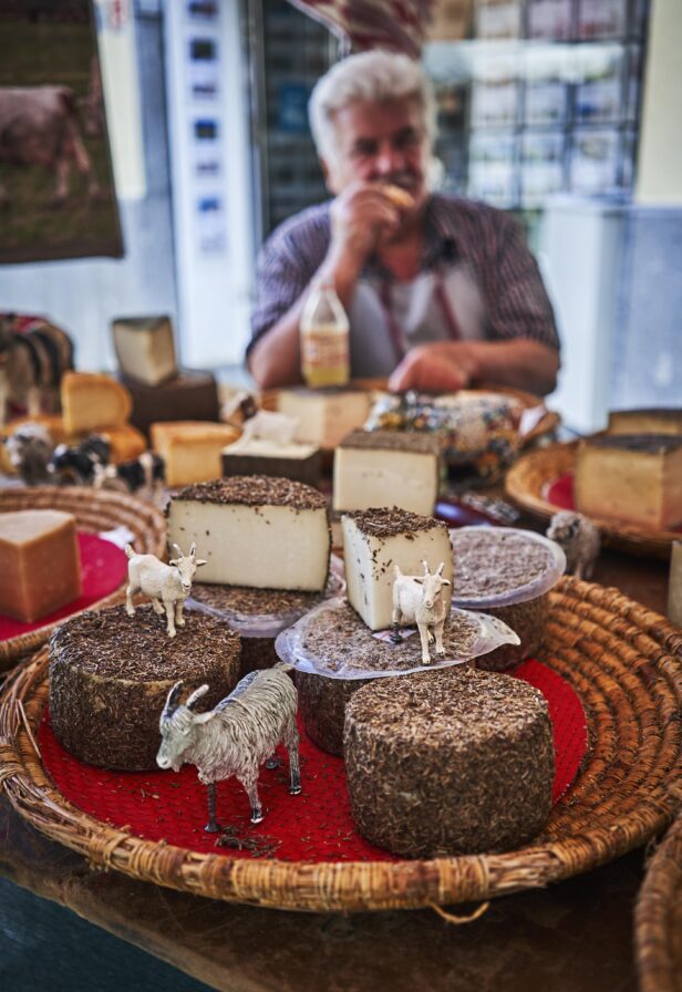 A variety of cheese wheels with herbs on a table, with small goat figurines placed on them. A person sits in the background holding a beverage.