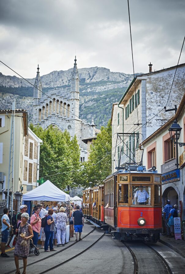 A vintage tram passes through a street market in a scenic village with mountains in the background and a church visible behind trees.
