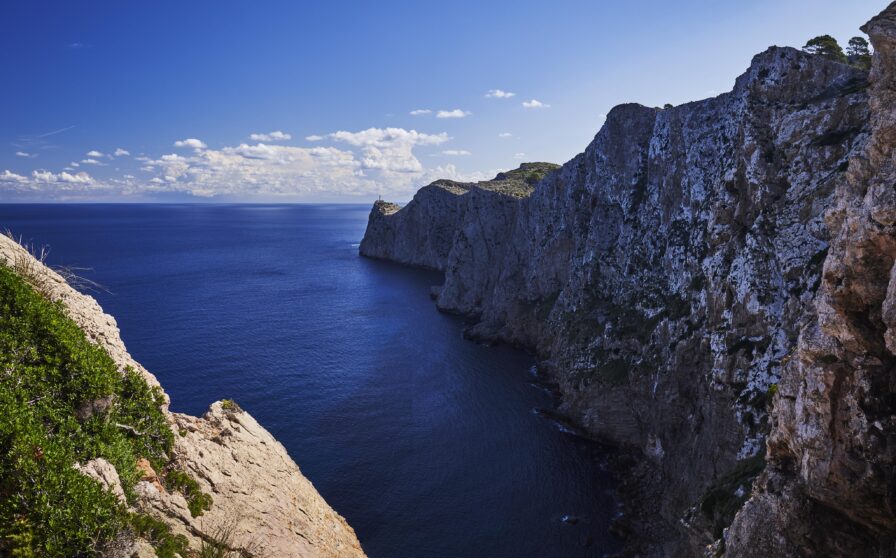 Steep rocky cliffs over a calm blue sea under a clear sky with scattered clouds. Sparse vegetation on cliff tops.