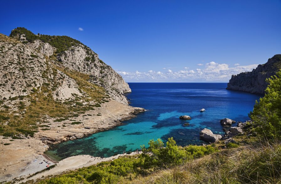 A scenic view of a rocky coastline with a small bay featuring clear turquoise water. A couple of boats are visible on the water, and greenery covers the surrounding cliffs under a bright blue sky.
