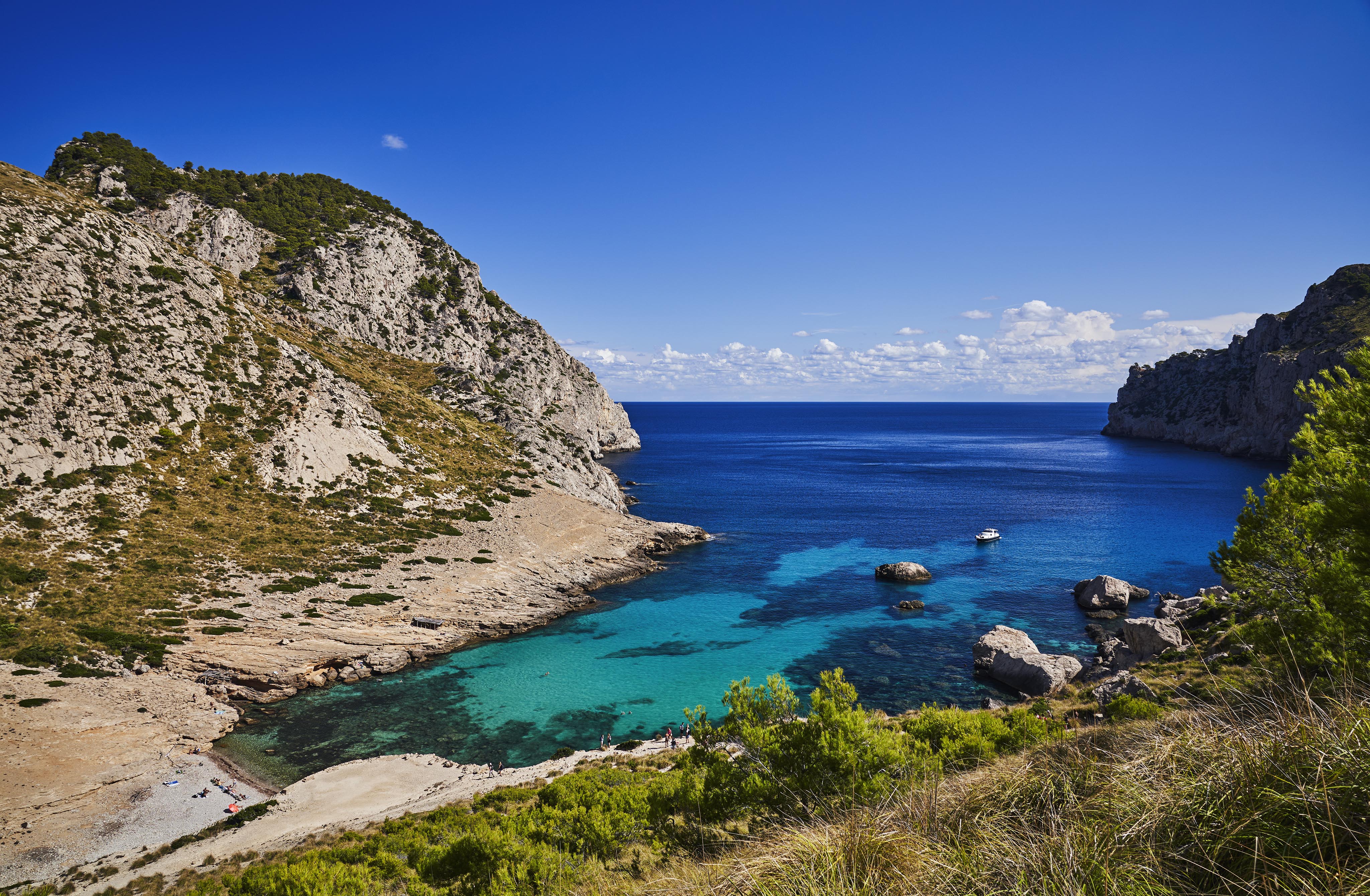 A scenic view of a rocky coastline with a small bay featuring clear turquoise water. A couple of boats are visible on the water, and greenery covers the surrounding cliffs under a bright blue sky.
