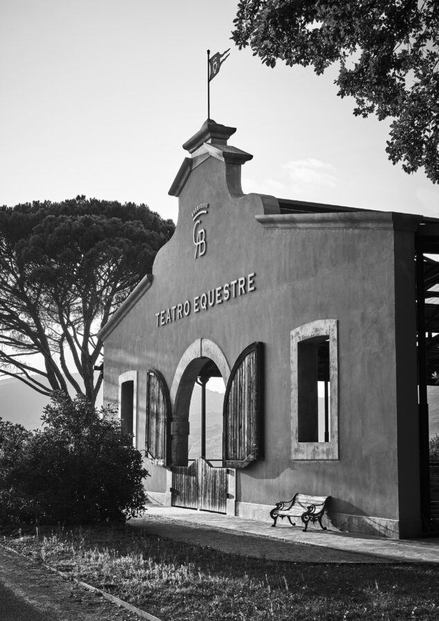 Black and white image of an Umbria equestrian theater building with arched entrances, a flag on top, and a nearby bench. Italian landscape in the background.