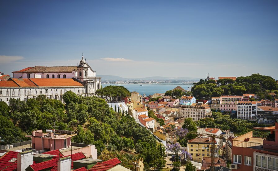A scenic view of Lisbon with historic buildings, red rooftops, and tree-covered hills overlooking the blue waters of the Tagus River under a clear sky.