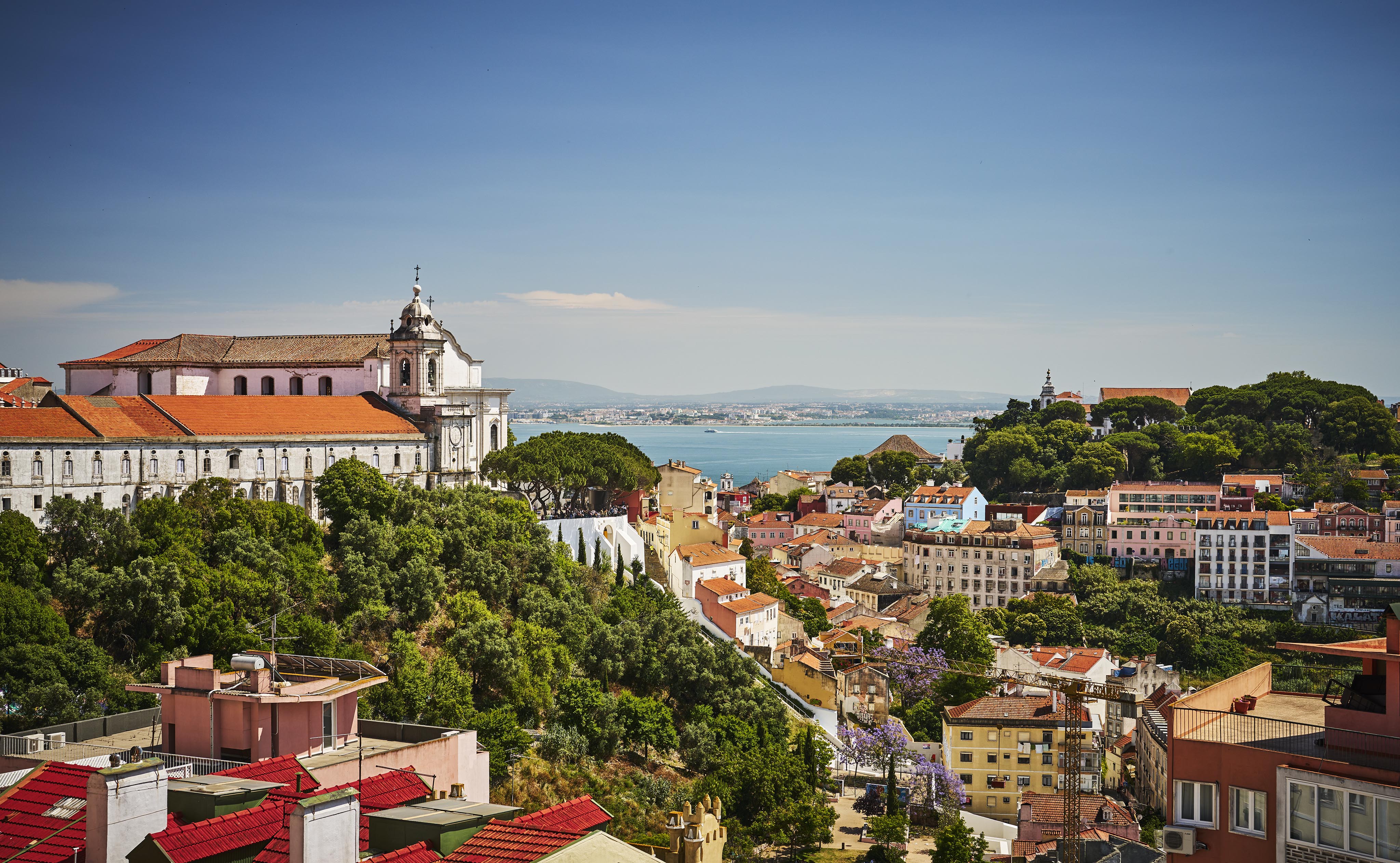 A scenic view of Lisbon with historic buildings, red rooftops, and tree-covered hills overlooking the blue waters of the Tagus River under a clear sky.