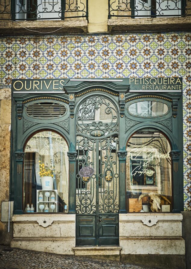 Vintage storefront with ornate metal door and decorative tiles. Signage reads "OURIVES PETISQUEIRA Restaurante". Two large windows display various items. Cobblestone sidewalk in front.