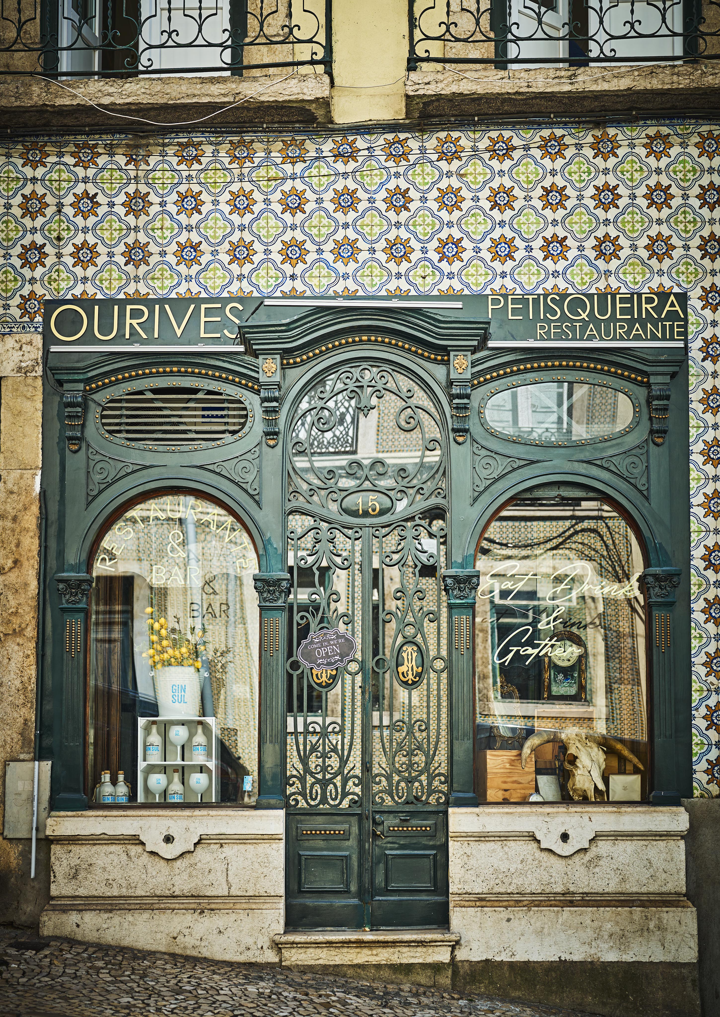 Vintage storefront with ornate metal door and decorative tiles. Signage reads "OURIVES PETISQUEIRA Restaurante". Two large windows display various items. Cobblestone sidewalk in front.