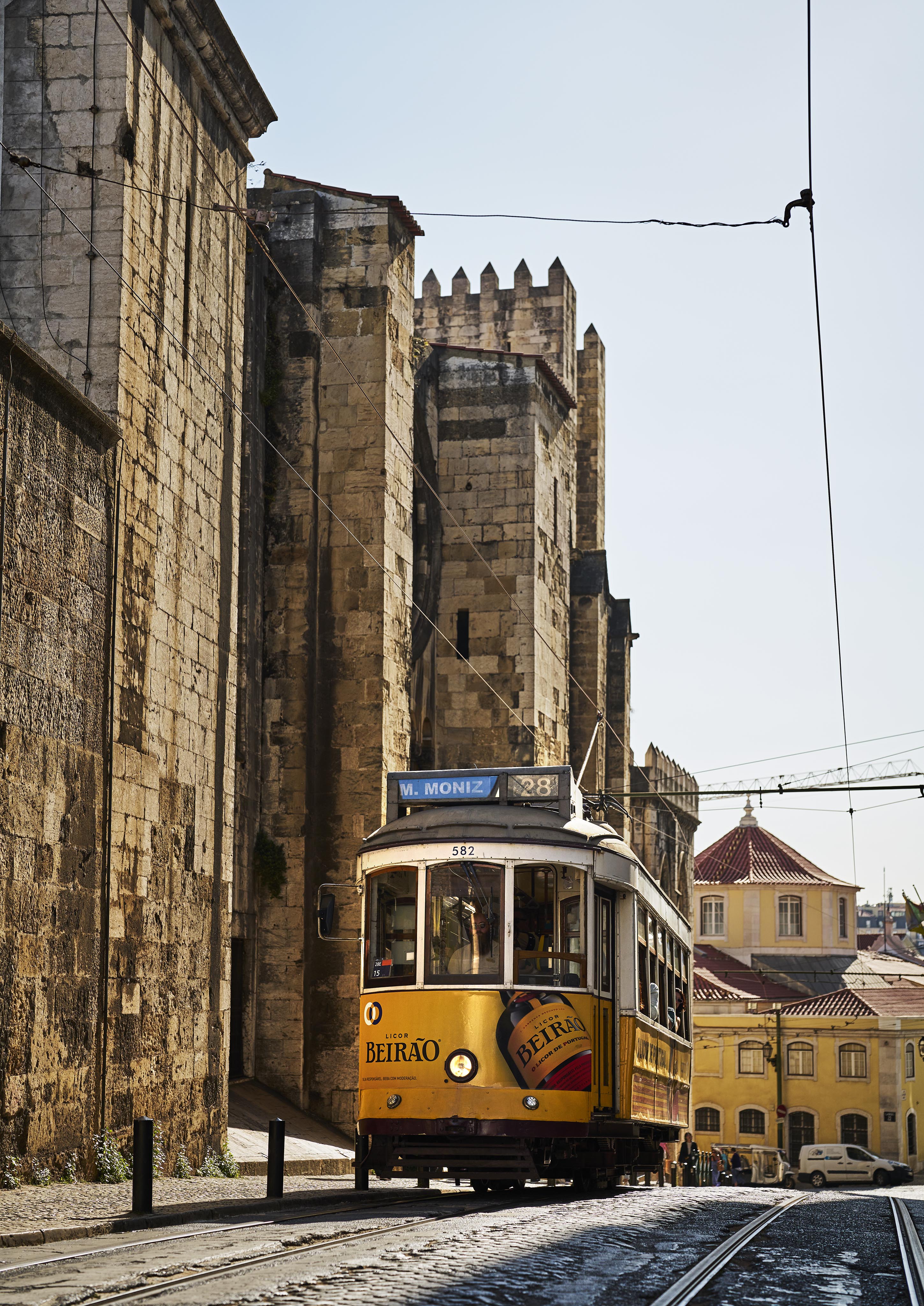 A vintage yellow tram travels along a street in front of a historic stone building with arches and battlements under a clear sky.