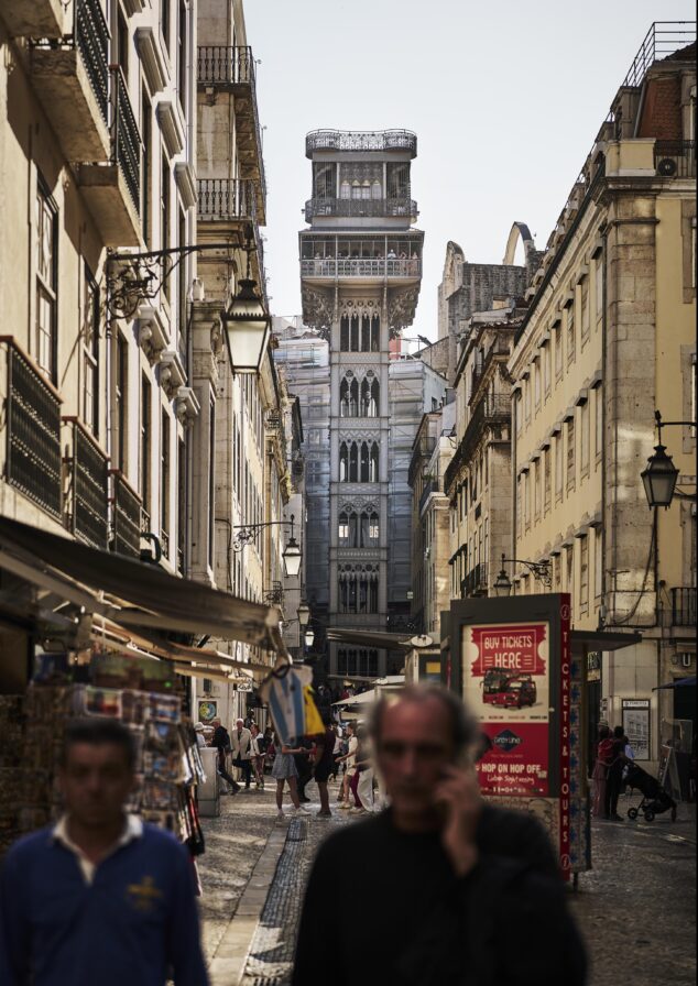 A narrow street leads to the Santa Justa Lift in Lisbon. People walk along the cobblestone path, flanked by buildings and street lamps.