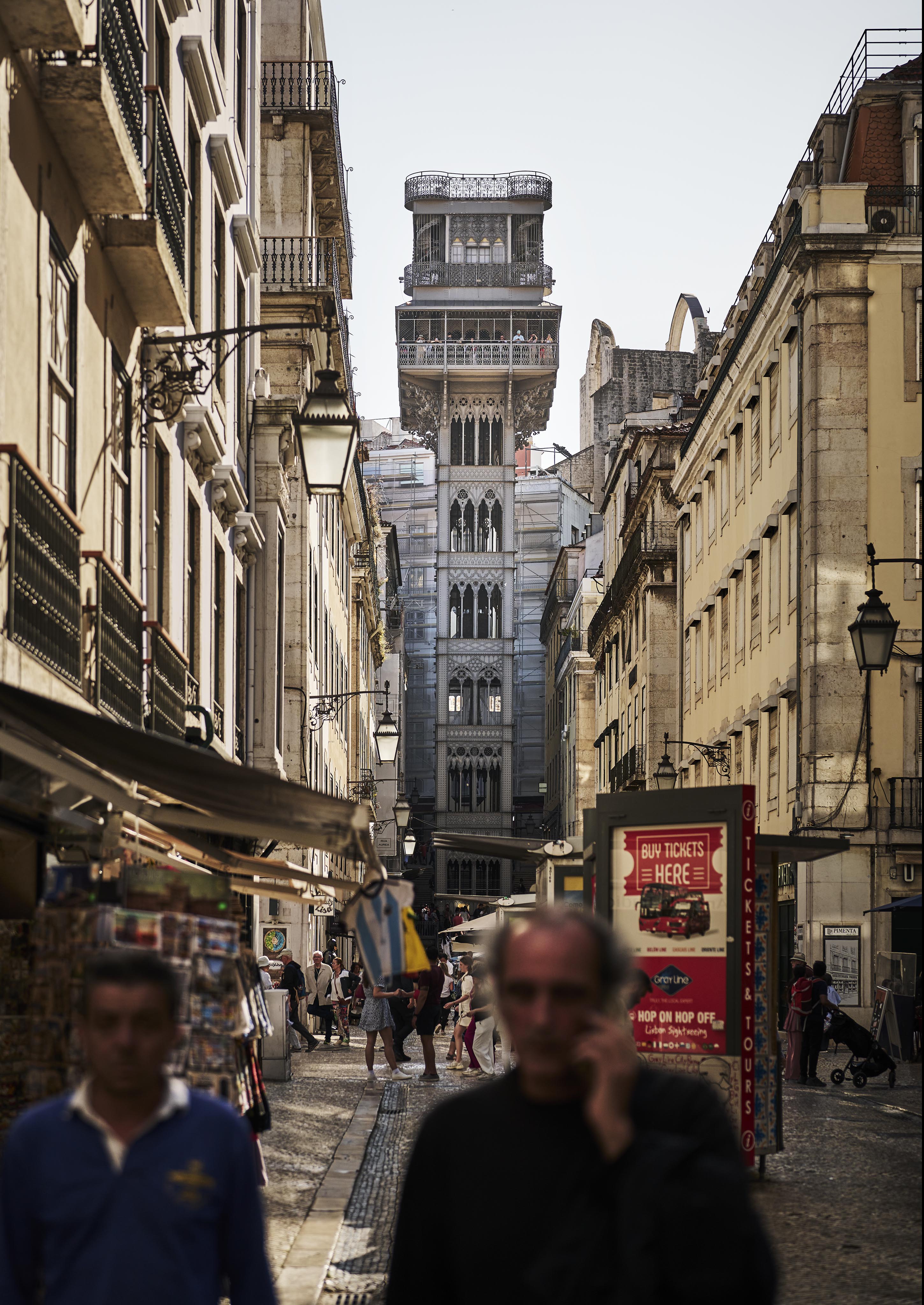 A narrow street leads to the Santa Justa Lift in Lisbon. People walk along the cobblestone path, flanked by buildings and street lamps.