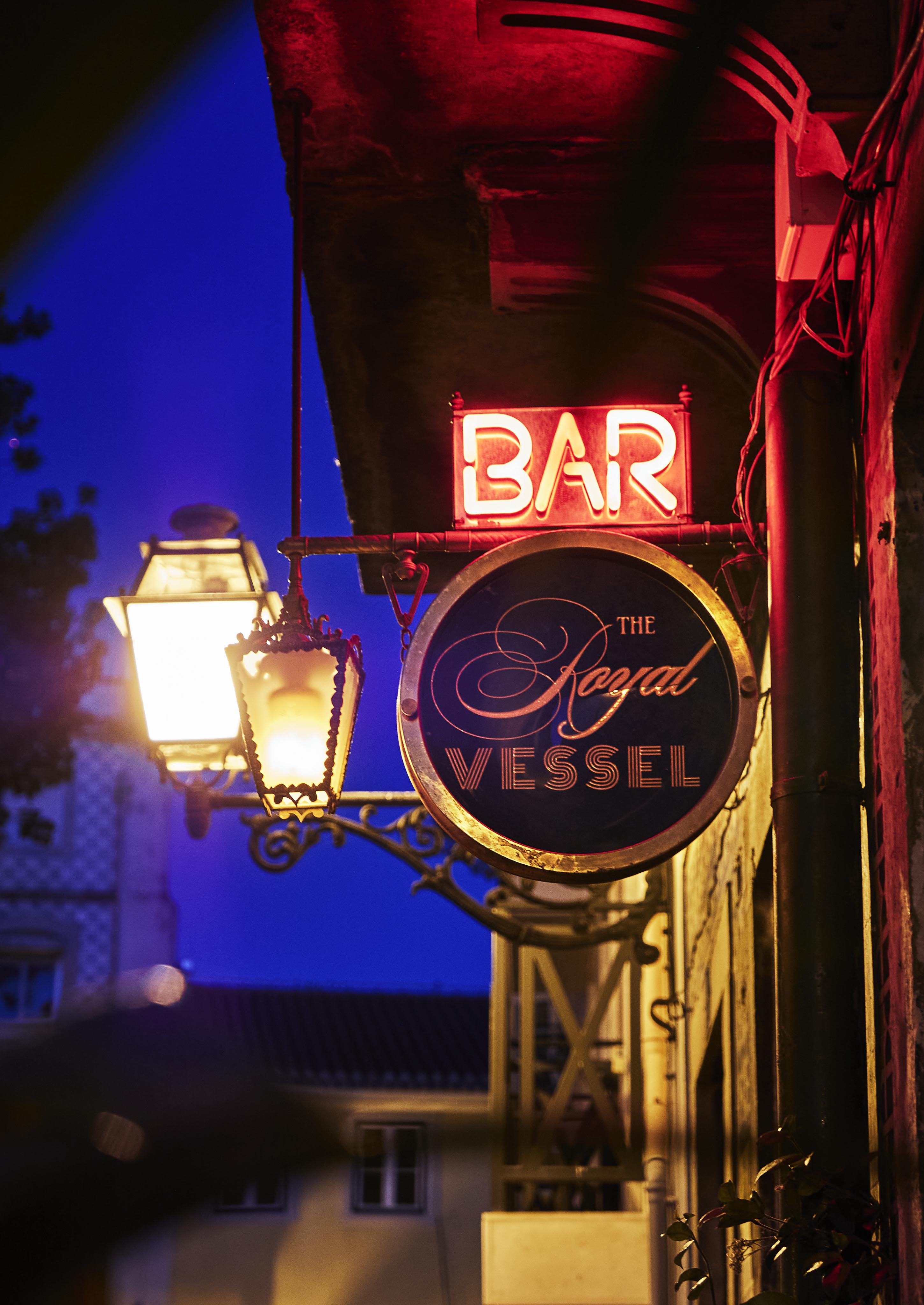 Neon sign for "The Royal Vessel" bar hangs under a streetlamp at dusk.