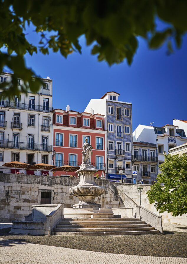 A stone statue stands on a fountain with steps in a sunny plaza, surrounded by colorful European-style buildings and trees.