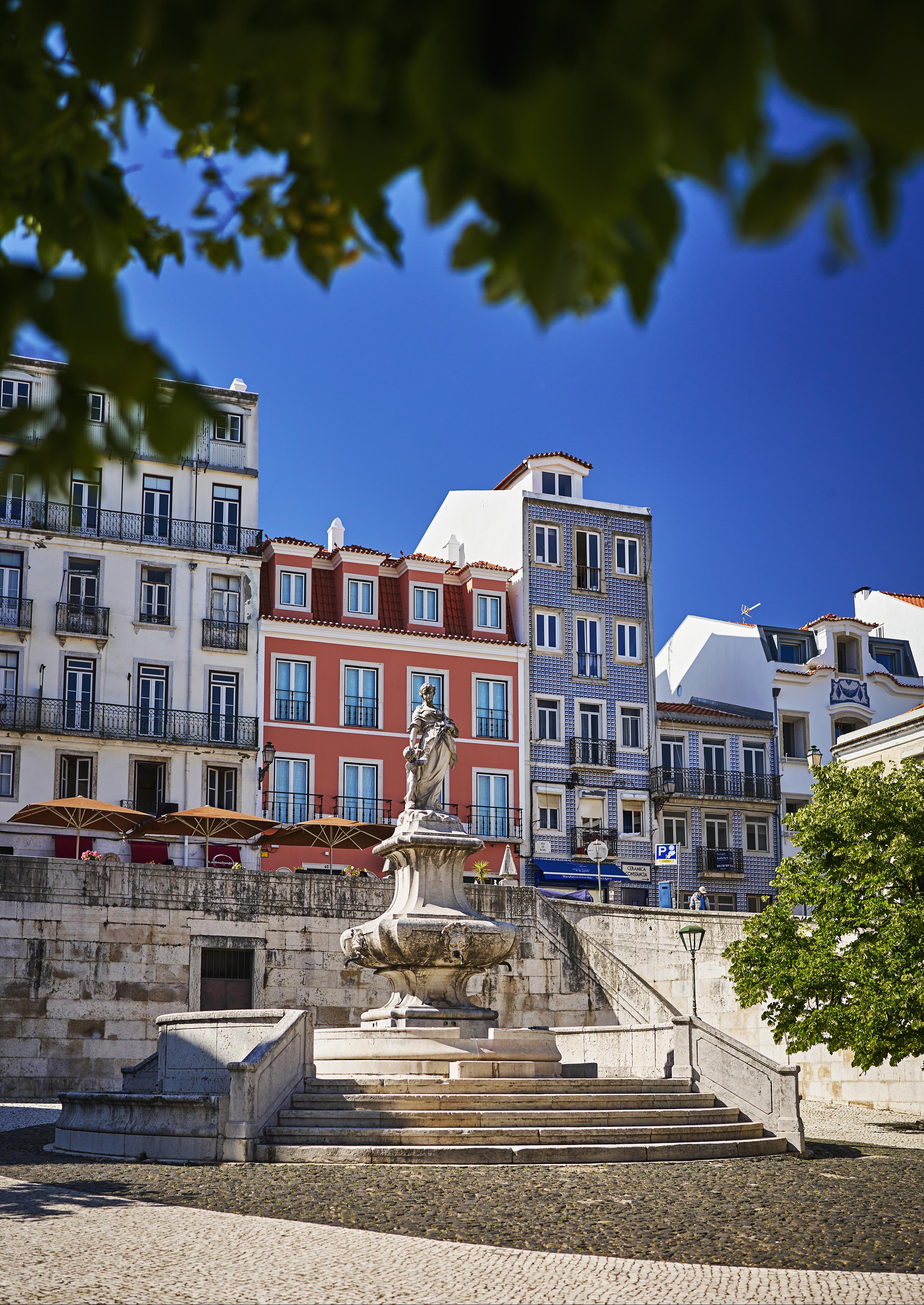 A stone statue stands on a fountain with steps in a sunny plaza, surrounded by colorful European-style buildings and trees.