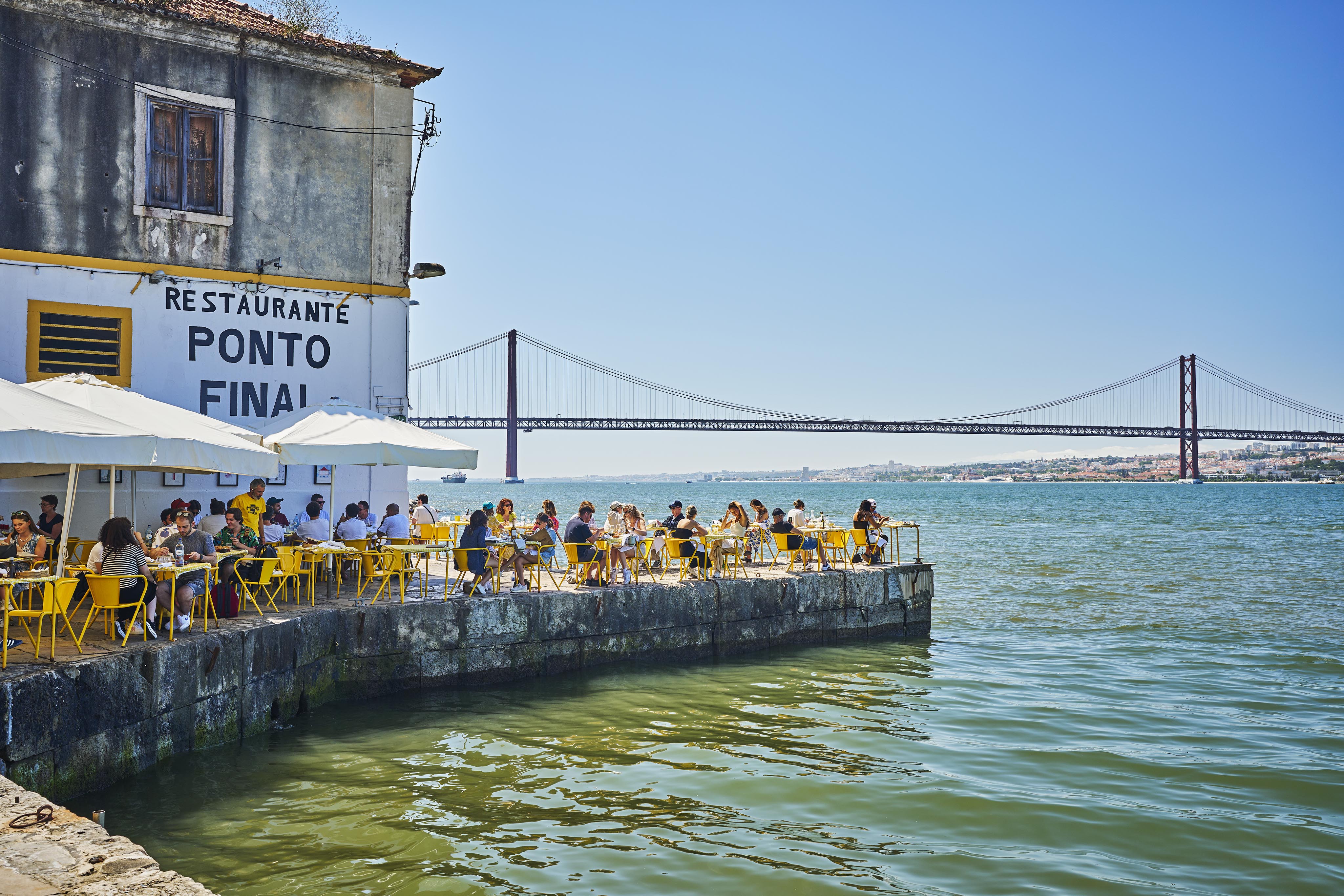 Outdoor diners sit at yellow tables by the water at Restaurante Ponto Final, with a large bridge and clear blue sky in the background.